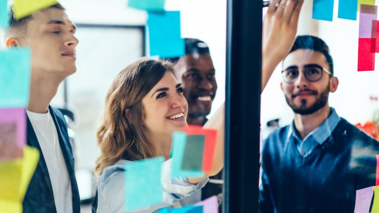 Fyra personer ser upp mot en glasvägg. En av dem sätter upp en lapp på väggen. Flera lappar i olika färger är redan uppsatta där.