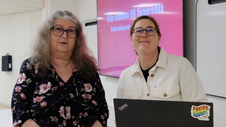 Christina har långt, grått hår, glasögon och blommig blus. Hanna har brunt, uppsatt hår, glasögon och vit skjorta. Båda småler mot kameran.