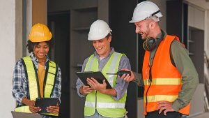 Tre byggarbetare i skyddsvästar och hjälm. Två håller i varsin läsplatta som de tittar på. Alla ler.
