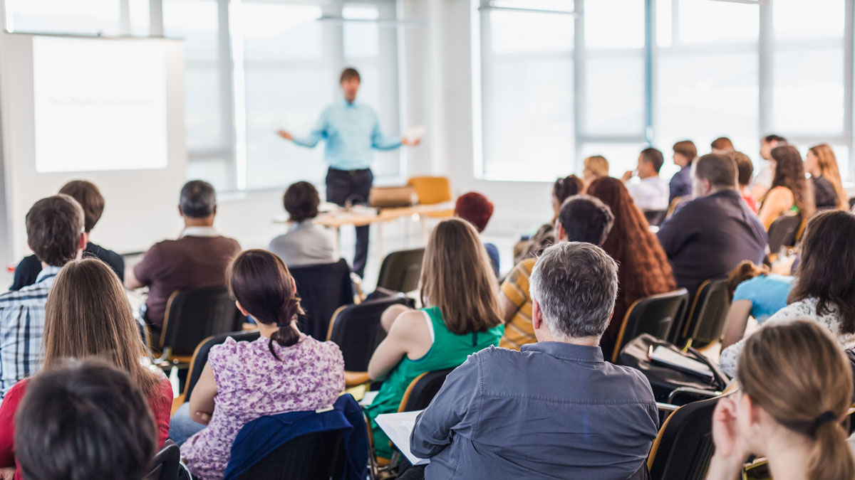 Flera personer sitter i en konferenssal och lyssnar på en talare längst fram framme i lokalen,