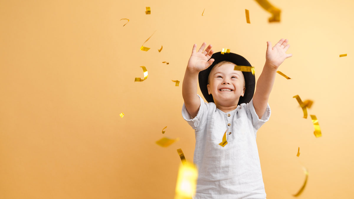 En ung pojke med Downs syndrom står i ett konfettiregn. Han ler stort och sträcker ut händerna för att fånga konfetti.