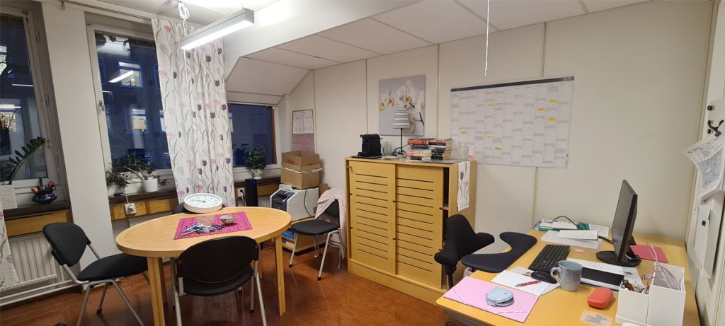 Två väggar i ett kontorsrum. Väggen till höger har tre fönster. I hörnet står en skrivare och intill den ryms en stol, en djup hylla och i hörnet ett skrivbord.