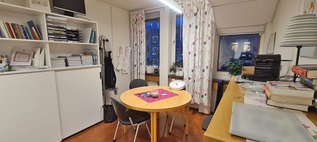 Tre väggar av ett rum. Till vänster en bred bokhylla, sedan lite tomt utrymme i hörnet. Mittenväggen har tre fönster, varav det till höger är lite mindre, eftersom taket är lägre precis där. På golvet ryms ett runt bord och längst vänsterväggen står en djup hylla.