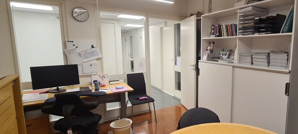 Ingången till ett kontorsrum; till vänster om dörren ryms ett skrivbord och en extrastol. Till höger avgränsas rummet av en vägg.