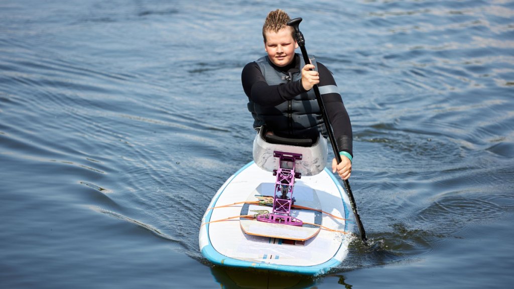 En ung kille sitter i en specialkonstruerad sits monterad på en paddelbräda, SUP. Han paddlar med en åra.