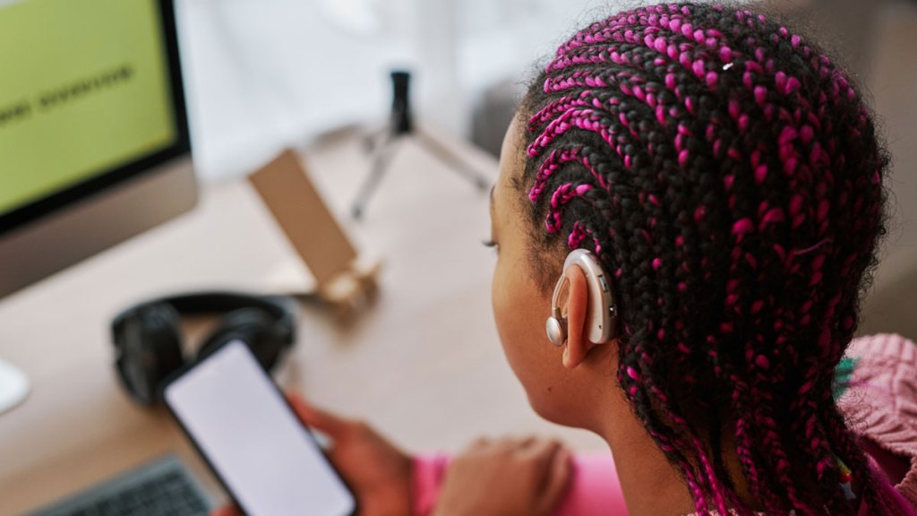 En ung tjej sitter framför en dator med en mobiltelefon i handen. Hon har en hörapparat.
