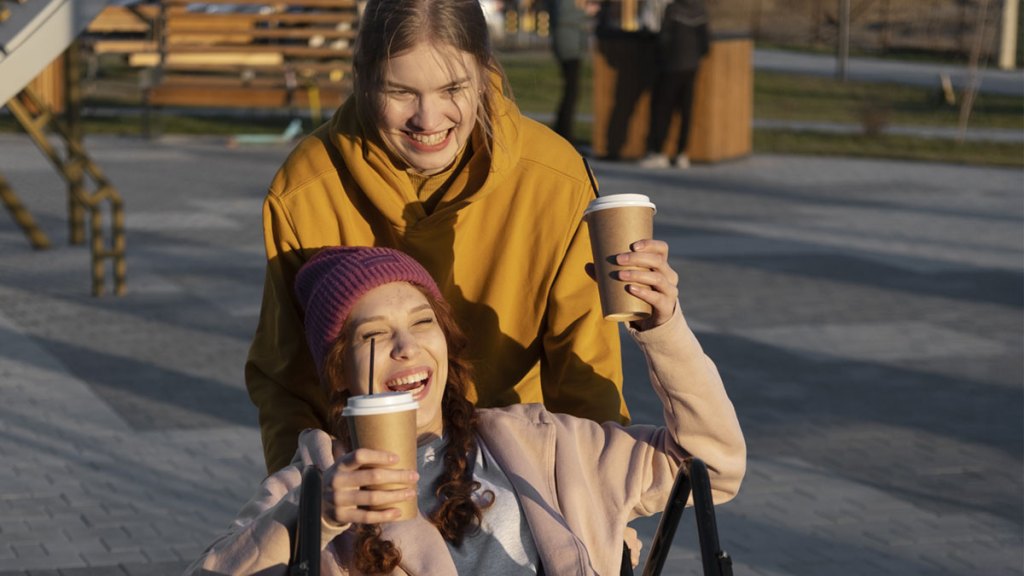 Två tonåringar som är utomhus. Den ena sitter i en rullstol och håller engångsmuggar i papp i båda händerna. Den andra kör rullstolen på bakhjulen. Båda skrattar.