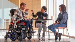 Tre personer sitter vid ett bord och har ett möte. En kille använder rullstol. De kommunicerar på teckenspråk.