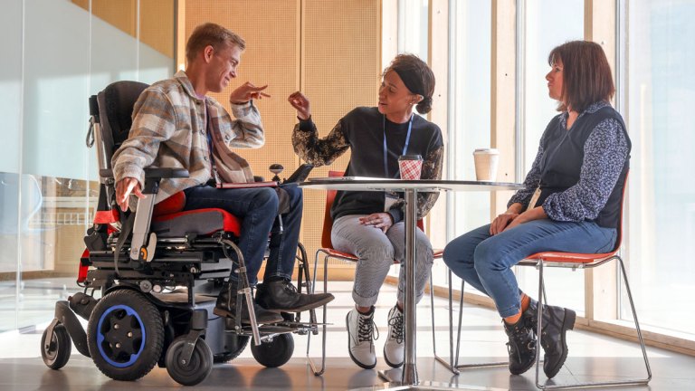 Tre personer sitter vid ett bord och har ett möte. En kille använder rullstol. De kommunicerar på teckenspråk.