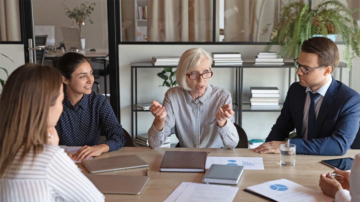 Ett möte. En kvinna med vitt hår och glasögon talar, medan de andra lyssnar uppmärksamt.