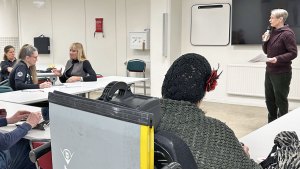 Generalsekreterare Sanna talar i en mikrofon. Runt henne i hästskoformering sitter deltagarna och lyssnar.