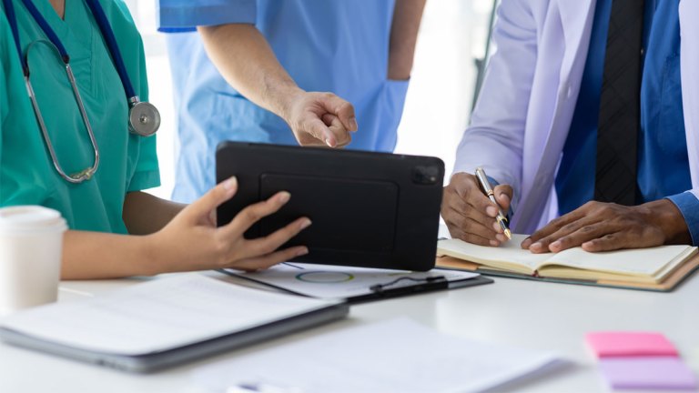 Två personer i sjukskötarkläder sitter vid ett bord och tittar på en platta som en av dem håller upp. En tredje person står mellan dem och pekar mot plattan.