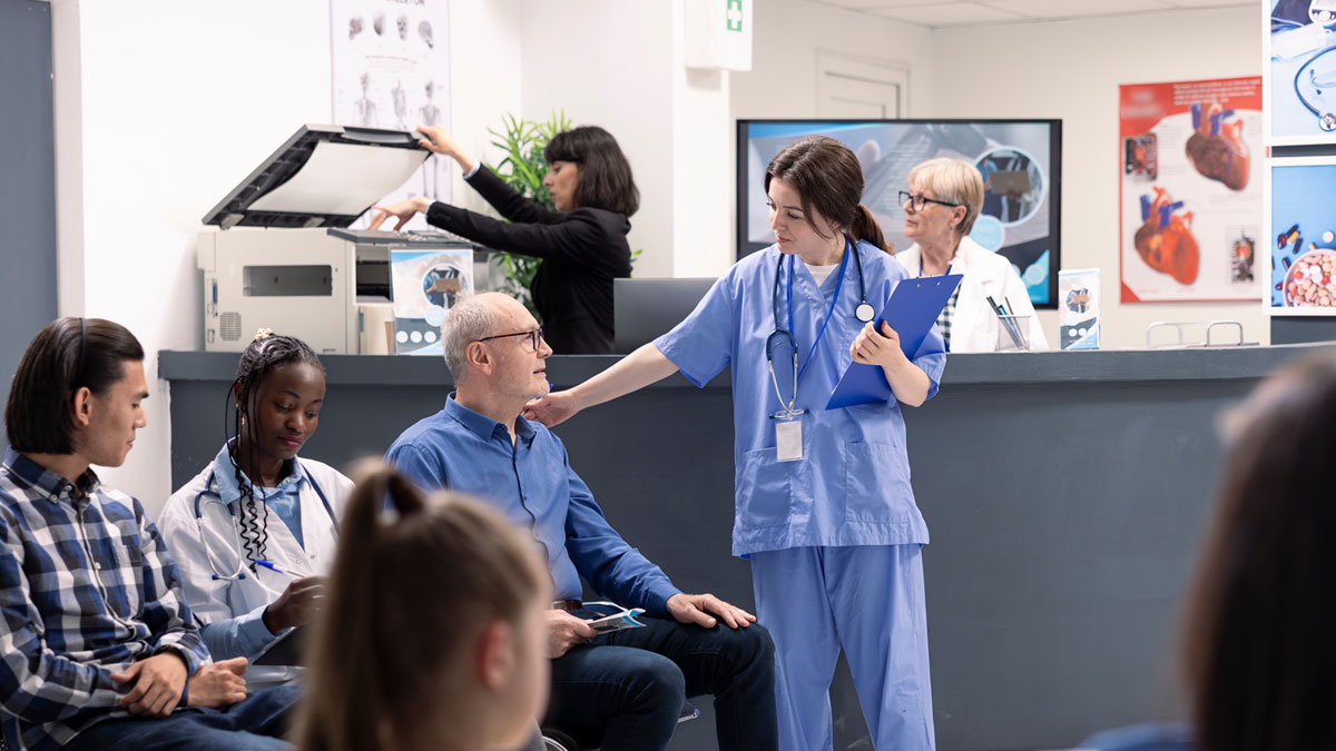 Flera personer sitter i ett väntrum på ett sjukhus. En sjuksköterska lägger en lugnande hand på en mans axel.