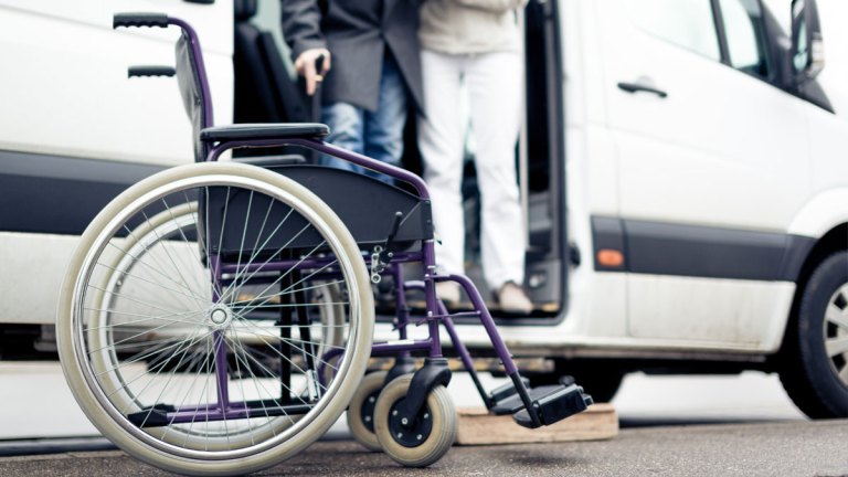 En sjuksköterska hjälper en man med käpp att ta sig ut ur en färdtjänstbil. I förgrunden står en rullstol.