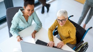 Två kvinnor sitter tillsammans vid ett bord. En sitter i rullstol och knappar på ett tangentbord. Den andra håller i en pappersbunt. Båda ser mot en datorskärm.