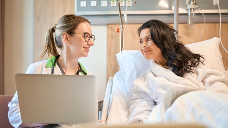 En läkare sitter vid en sjukhussäng. Hon håller en laptop. I sängen ligger en patient med slang i näsan. De ser på varandra och ser nöjda ut.