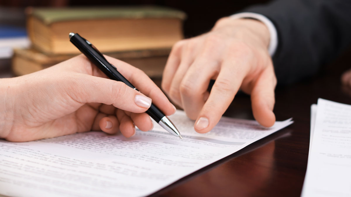 En hand pekar på ett dokument som en annan persons hand är på väg att skriva på.