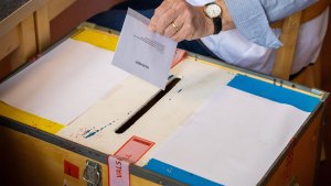 En hand håller ett valkuvert över en stor låda som är både låst och förseglad med tejp. lådan har en skåra på ovansidan, där kuvertet kan läggas ner.