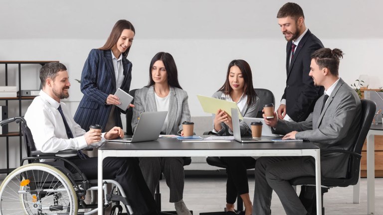 En grupp tjänstepersoner samlade kring ett mötesbord. En sitter i rullstol. De flesta har en kaffemugg.