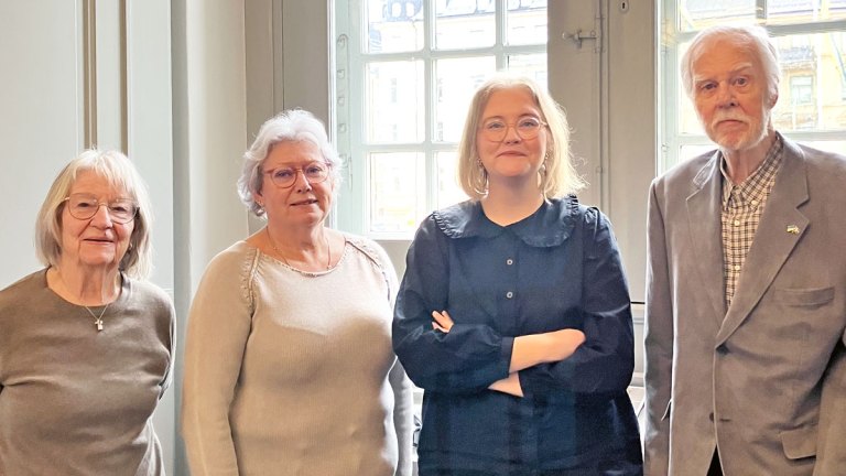 Fyra personer står på rad framför ett fönster och ler mot kameran.