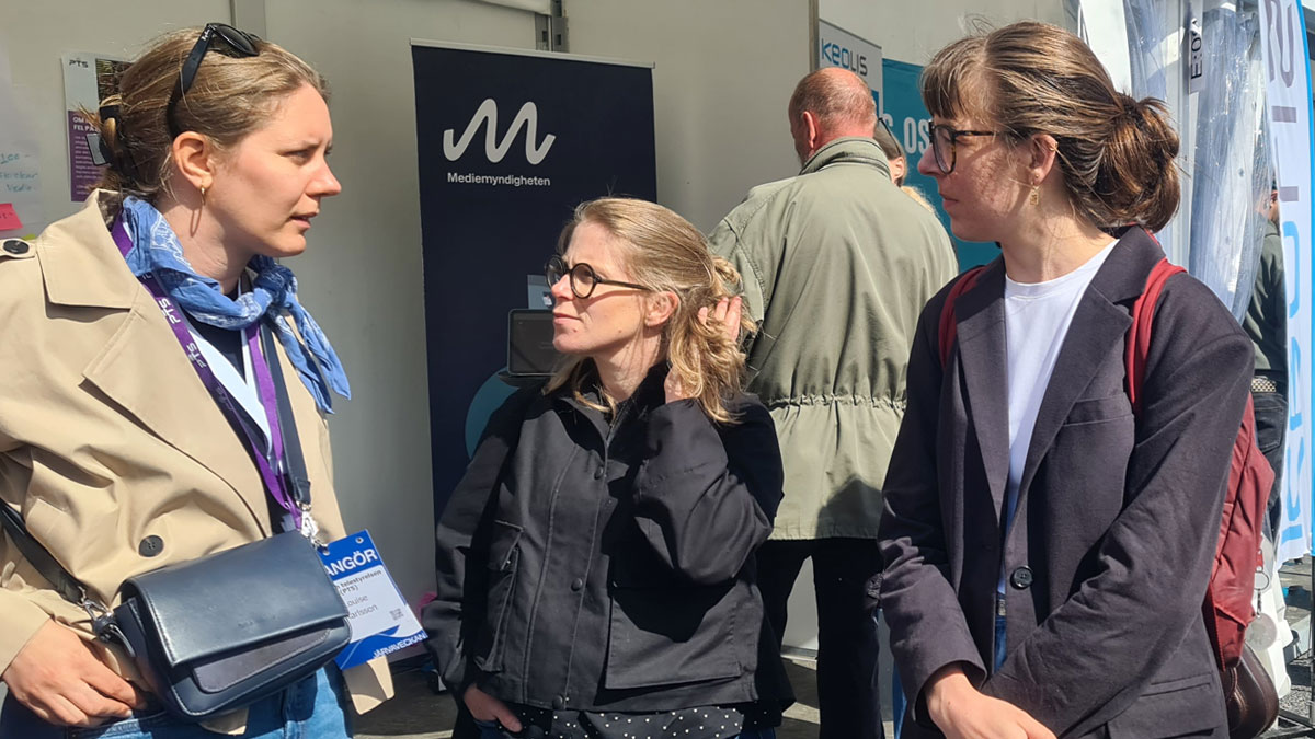 En mörkblond kvinna med en arrangörsbricka runt halsen säger något till två kvinnor, båda med glasögon, som uppmärksamt ser på henne.
