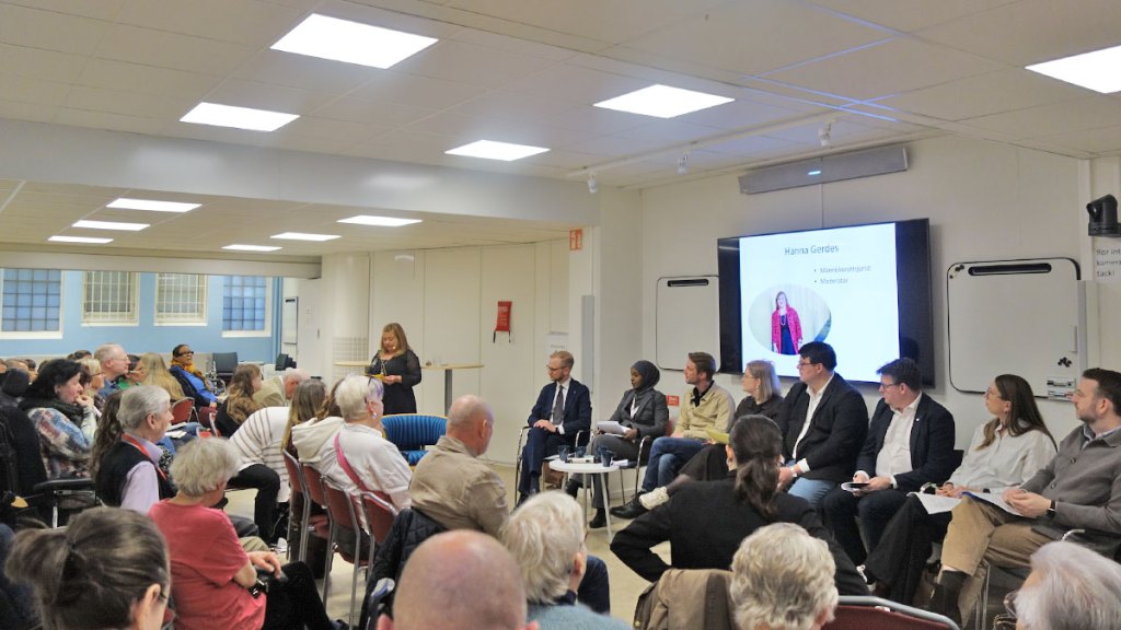 Konferenssalen hos Funktionsrätt Stockholms län är fullsatt. I center av bilden står moderatorn Hanna Gerdes och pratar. Bakom henne sitter  politikerpanelen på rad. 