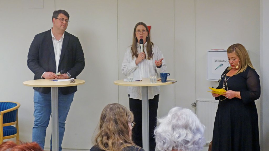 Msciwoj Swigon (SD) och Märta står vid varsitt högt, runt bord. Märta talar i  en mikrofon. Moderator Hanna Gerdes står intill och kikar i sina talkort.