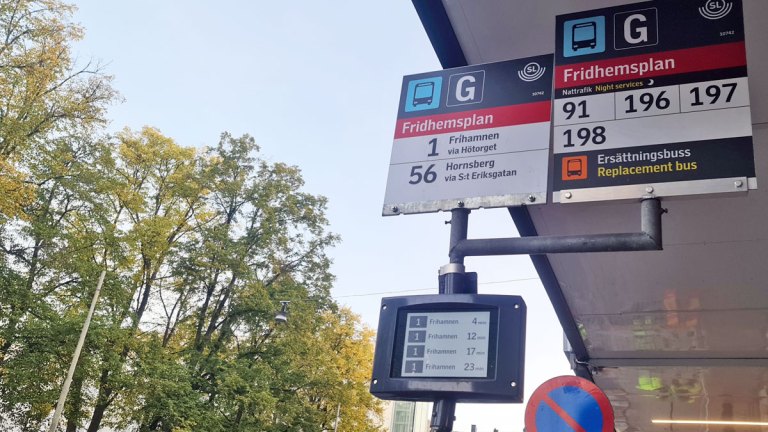 Två analoga skyltar vid en busshållplats informerar om vilka linjer som stannar där. En digital skylt under dem listar fyra bussar med beräknad ankomsttid.